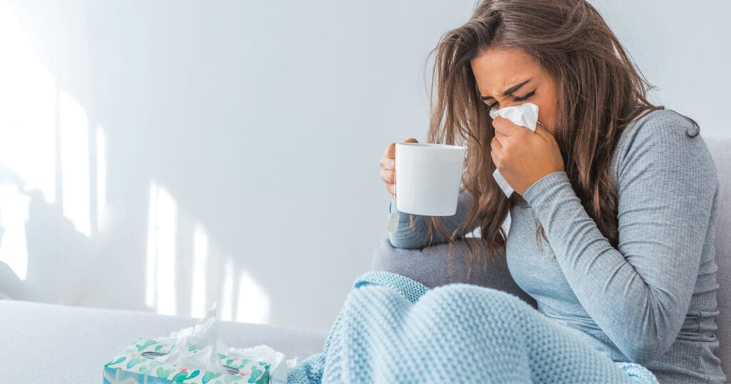 A young woman holds a mug while covering her face with a kleenex