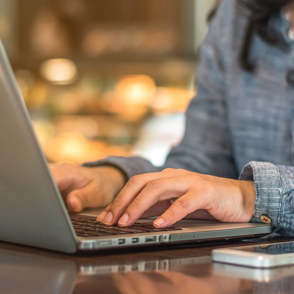 In a cozy cafe, a person types diligently on their laptop, crafting emails while a smartphone lies close at hand, ready to assist with paying bills seamlessly.