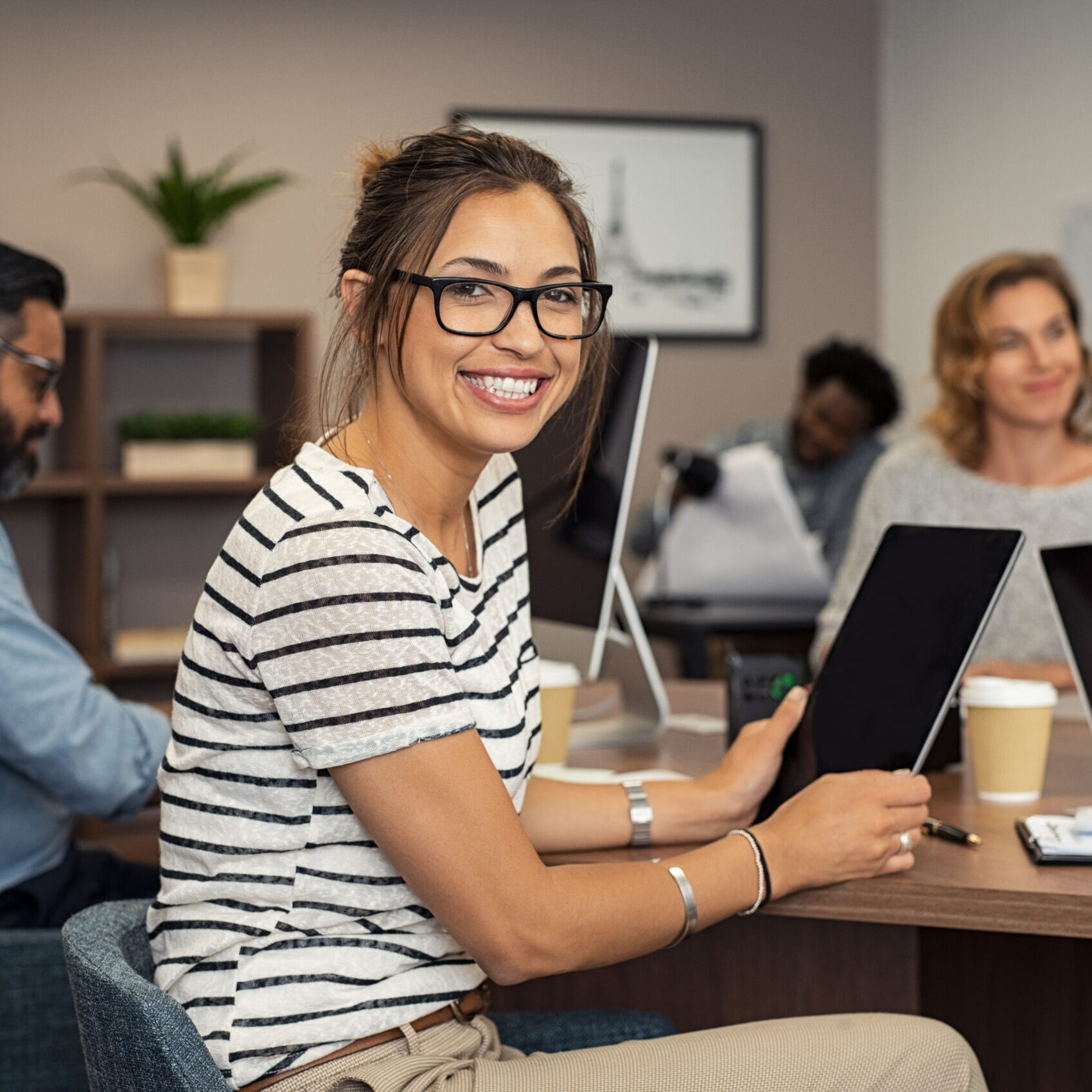 A person in glasses and a striped shirt sits at a table with a tablet, smiling, while others work with laptops and papers in the background, focusing on SEO strategies to enhance their employer services.