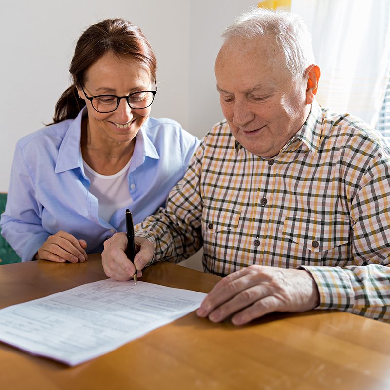 An elderly man signs documents at a table with a woman's assistance, both smiling, highlighting the supportive spirit of the Resident Resource Fair.